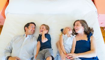 A family of four is lying on a bed, smiling and enjoying each other's company.