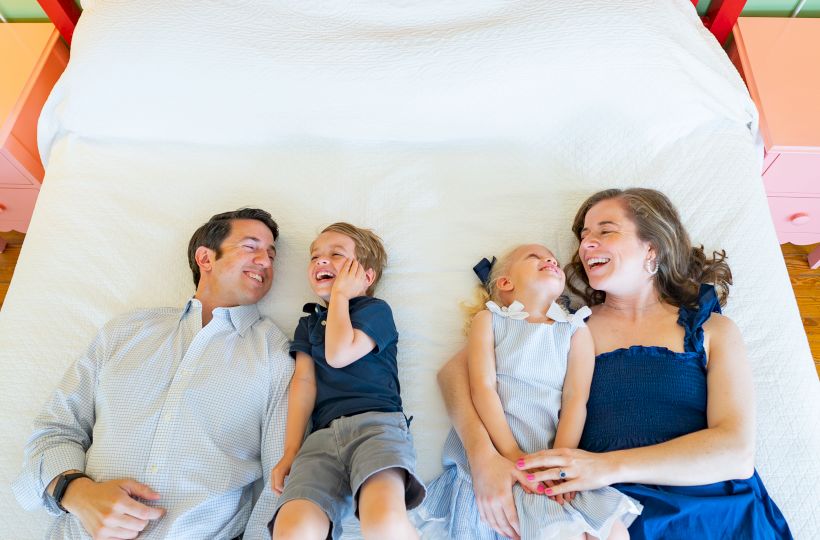A family of four is lying on a bed, smiling and enjoying each other's company.