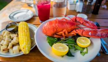 A lobster meal with lemon, corn on the cob, potatoes, a drink, and some greenery on a wooden table outdoors.