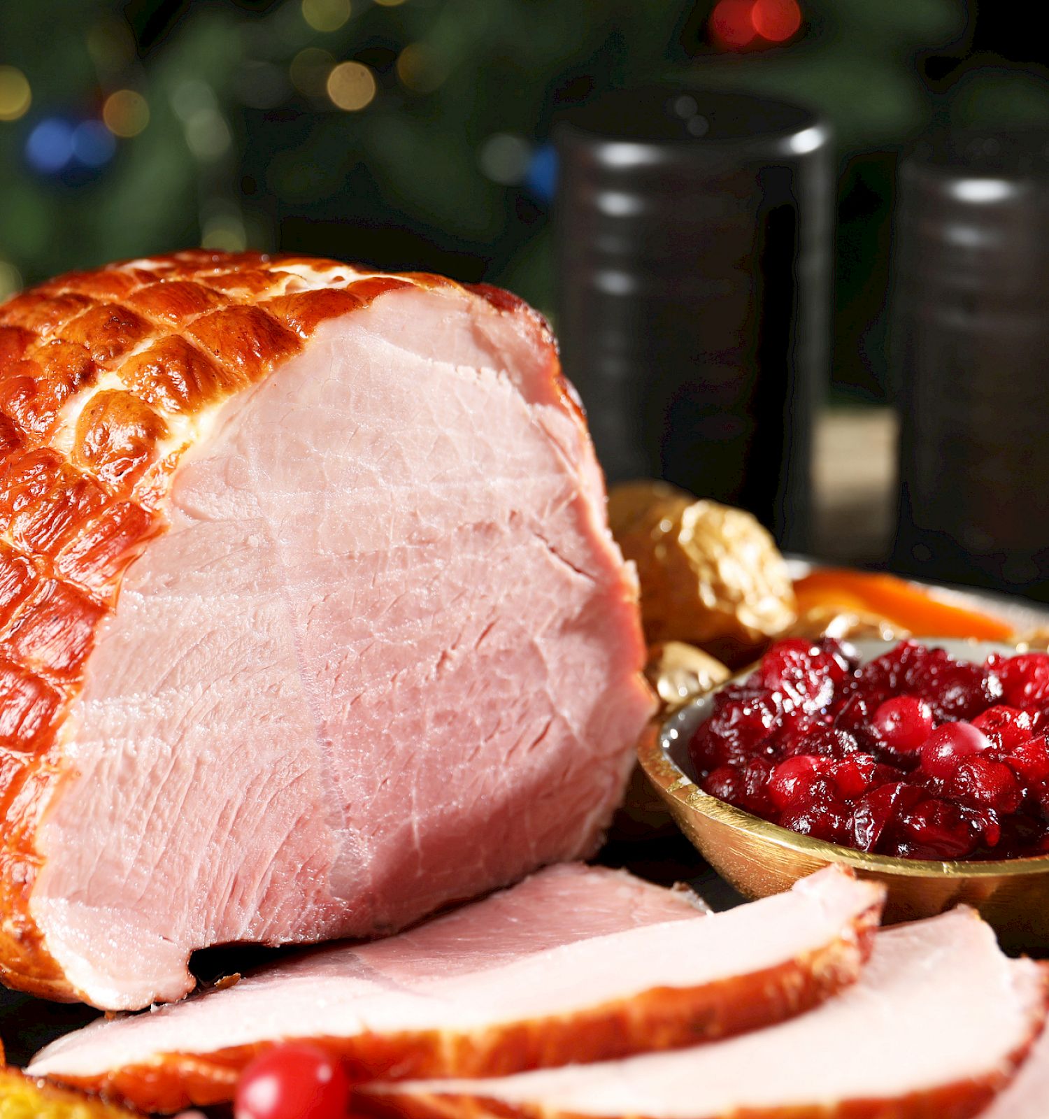 A roasted ham centerpiece on a festive table, sliced to reveal pink interior, surrounded by cranberry relish, oranges, and candles for a holiday feast.
