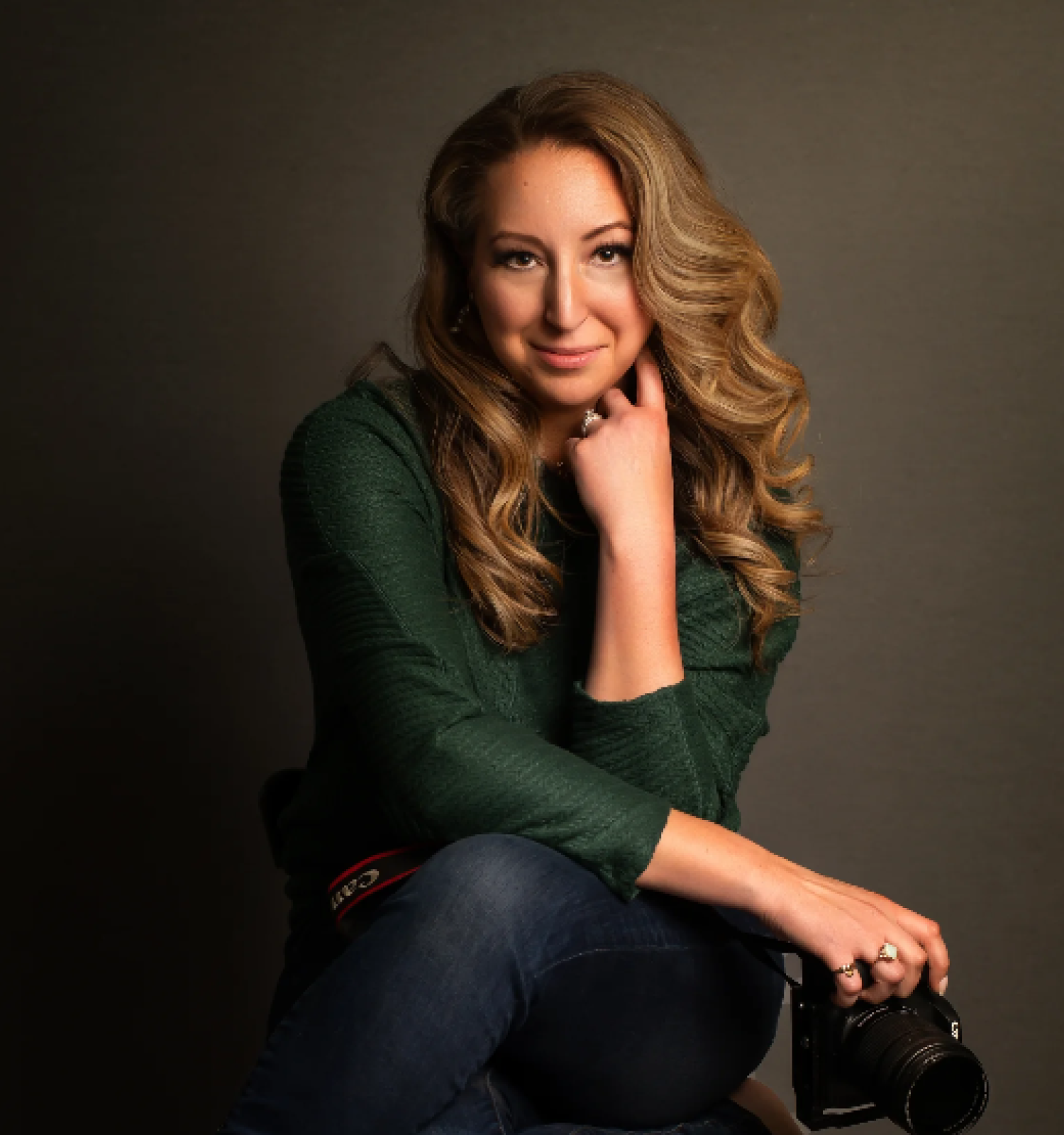 A woman with wavy blonde hair sits with a camera, wearing a green top and jeans, posing thoughtfully against a dark backdrop.