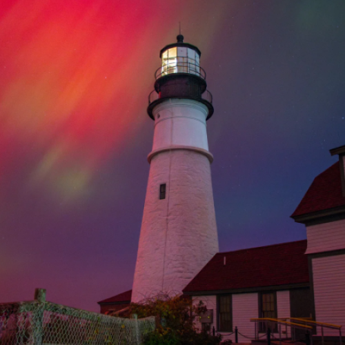 Photograph by Jennifer Egan featuring: A tall white lighthouse stands beside red-roofed buildings, under a dramatic night sky painted with vibrant, swirling auroras.