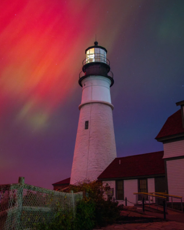 Photograph by Jennifer Egan featuring: A tall white lighthouse stands beside red-roofed buildings, under a dramatic night sky painted with vibrant, swirling auroras.