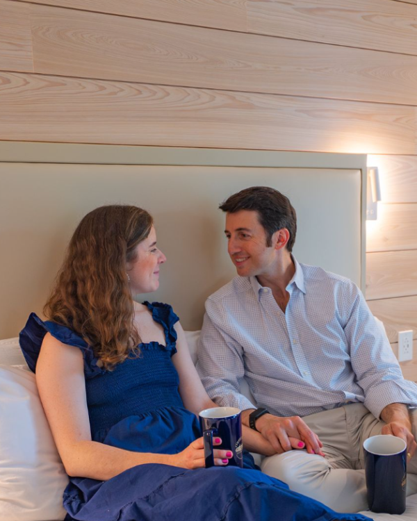 A couple sits on a bed, smiling and talking, enjoying drinks together in a cozy, wooden-paneled hotel room.