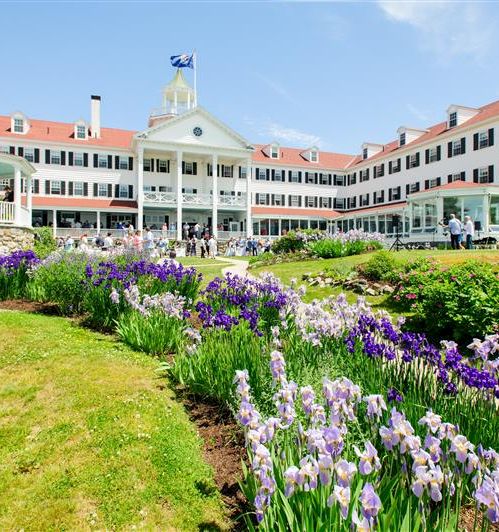 A grand white hotel with red roofs sits behind a colorful garden of purple and white flowers, sunny sky above, inviting front lawn.
