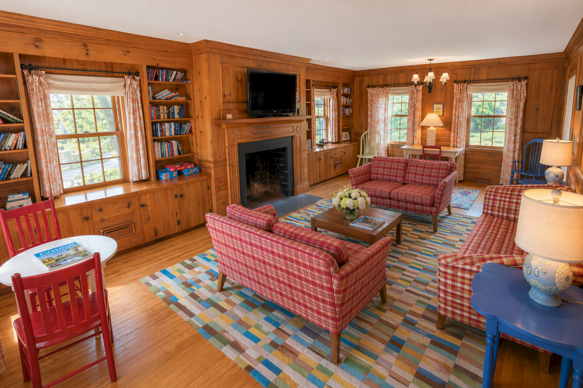 A cozy wood-paneled living room with plaid red sofas, a fireplace, built-in bookshelves, a blue side table, and a colorful rug under a central coffee table, sunny windows.
