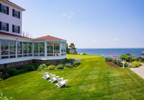 A seaside hotel lawn with a white-framed villa, glass-walled porch, green lawn, lounge chairs, and a view of the ocean under a sunny sky.