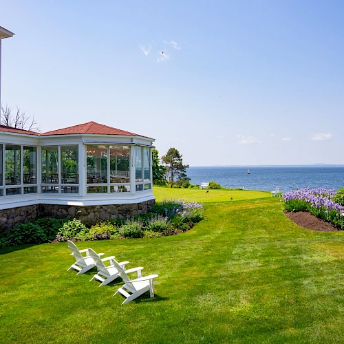 A seaside hotel lawn with a white-framed villa, glass-walled porch, green lawn, lounge chairs, and a view of the ocean under a sunny sky.