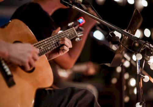 Two musicians playing acoustic guitars on stage, close-up of hands on strings, microphone and bokeh lights in the background, live performance ambience.