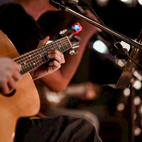 Two musicians playing acoustic guitars on stage, close-up of hands on strings, microphone and bokeh lights in the background, live performance ambience.