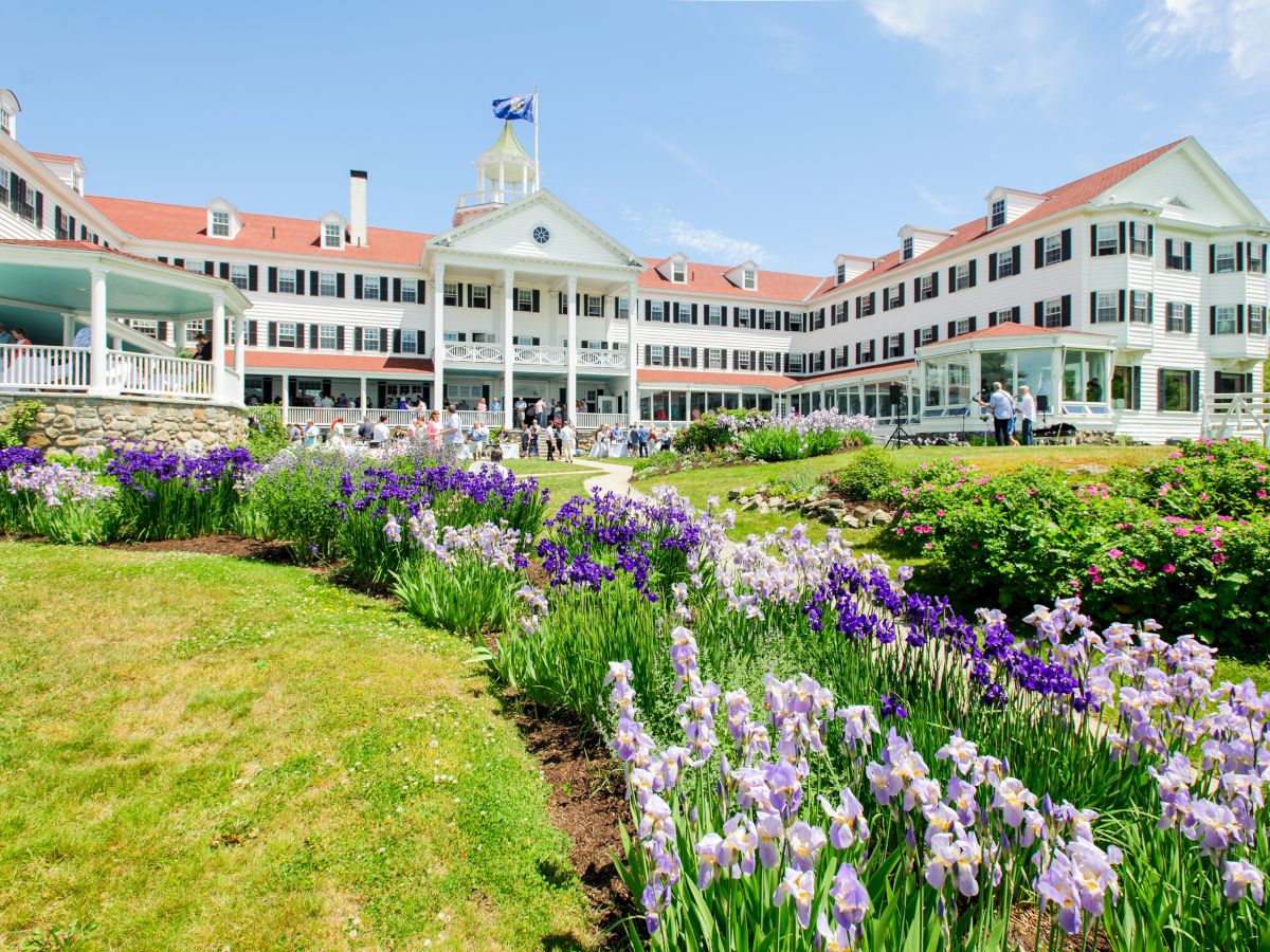 A sunny garden with blooming flowers in front of a large colonial-style school building where people gather.