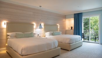 Bright modern hotel bedroom with two neatly made beds, wood-paneled wall, pendant lights, and a blue-curtained balcony door.