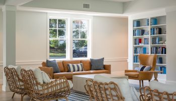 A bright living room with a tan sofa, rattan chairs, striped cushions, large windows, and built-in shelves of books.