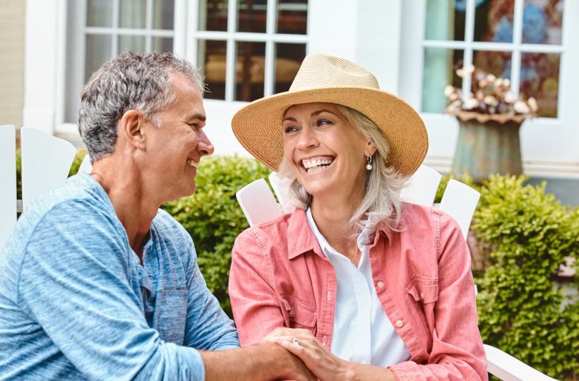 A couple sits on outdoor chairs, smiling at each other. The man wears a blue shirt, the woman a pink shirt and straw hat. Bushes are in the background.