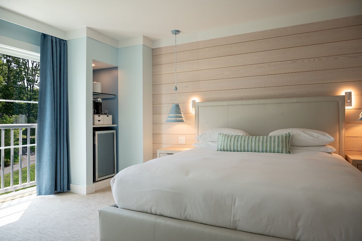 A modern bedroom with a large bed, layered wood wall paneling, pendant lamp, bedside lights, and blue curtains by a window.