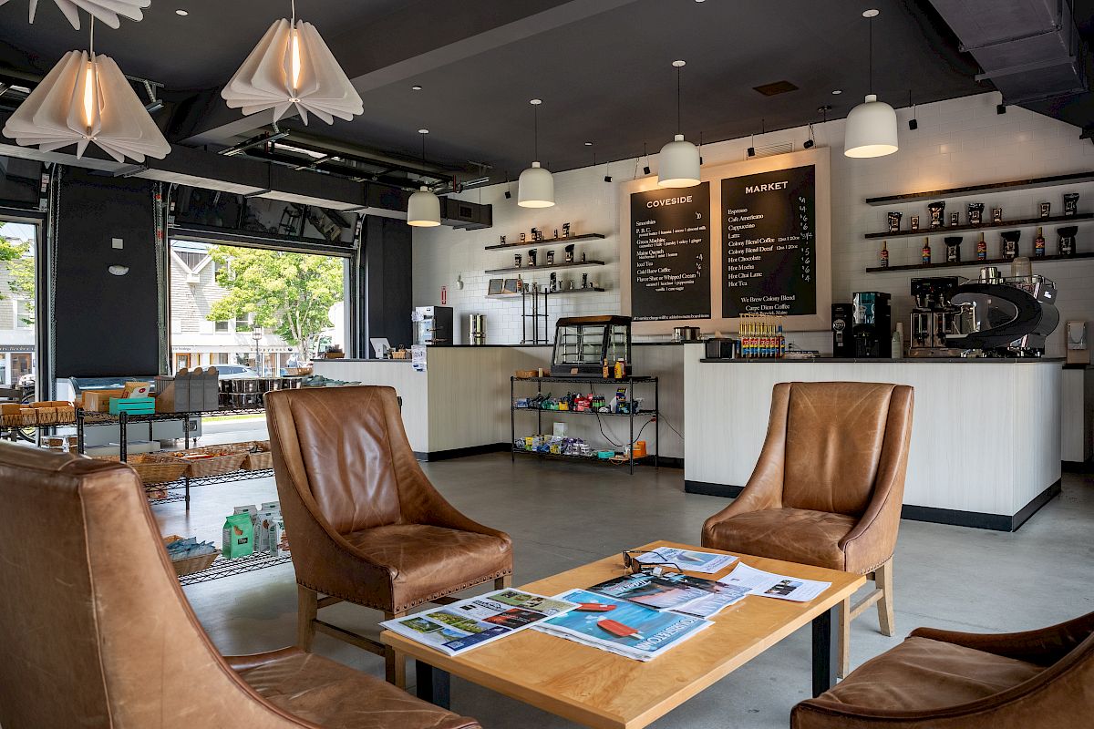 A modern coffee shop counter with an espresso machine, menu boards, drink shelves, and leather seating in the foreground.
