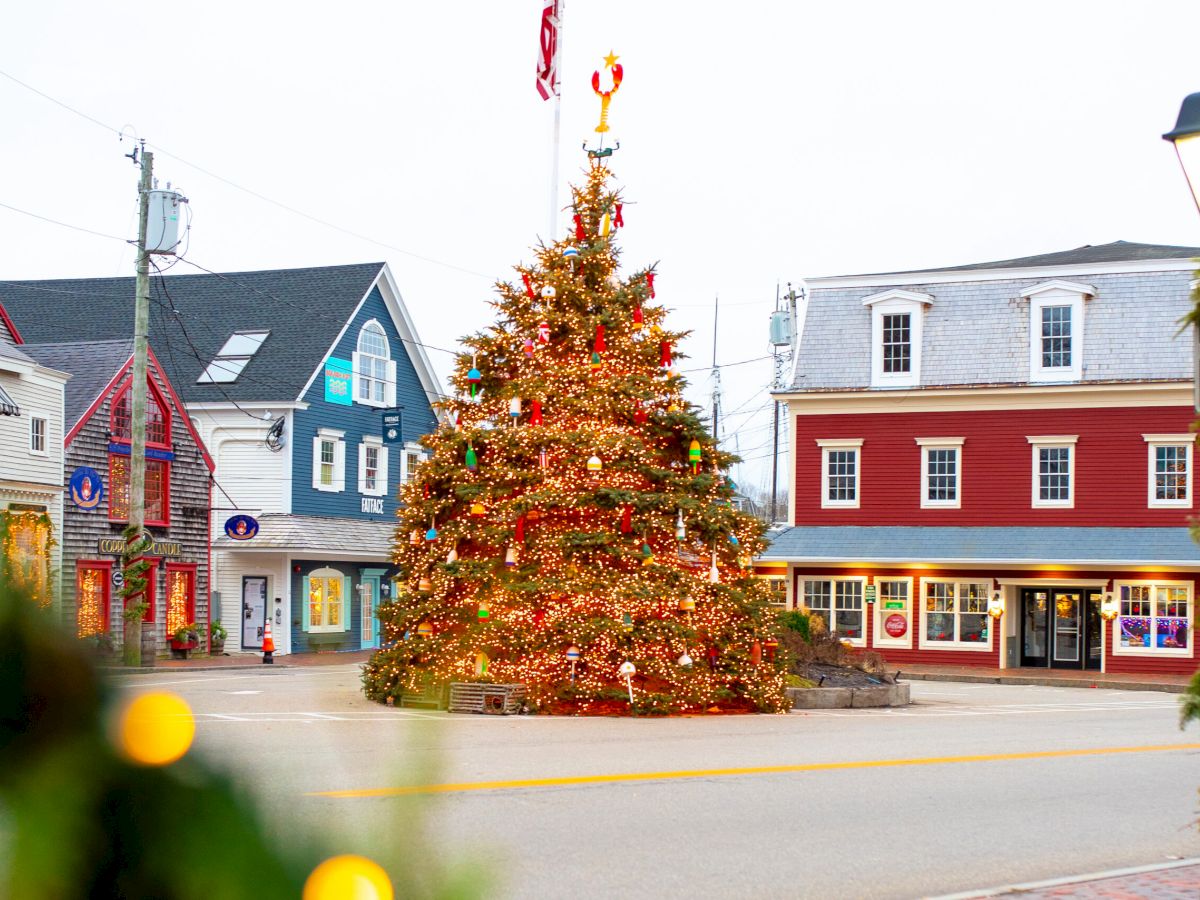 A large decorated Christmas tree glows in a town square beside storefronts and street lamps, with holiday lights everywhere.