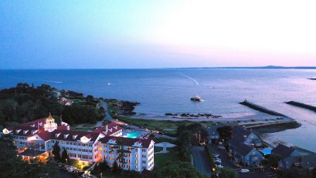 A seaside resort overlooks a calm bay with piers and boats at dusk, with lights glowing on white buildings.