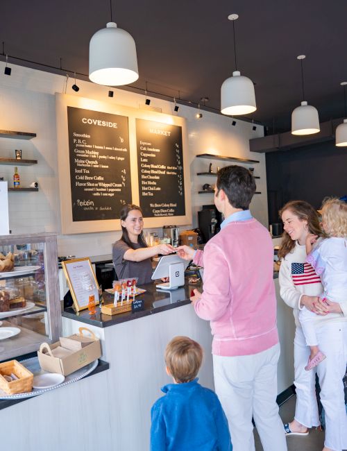 A cafe counter with hanging lights and menu boards, where a barista serves customers in line for drinks and snacks.